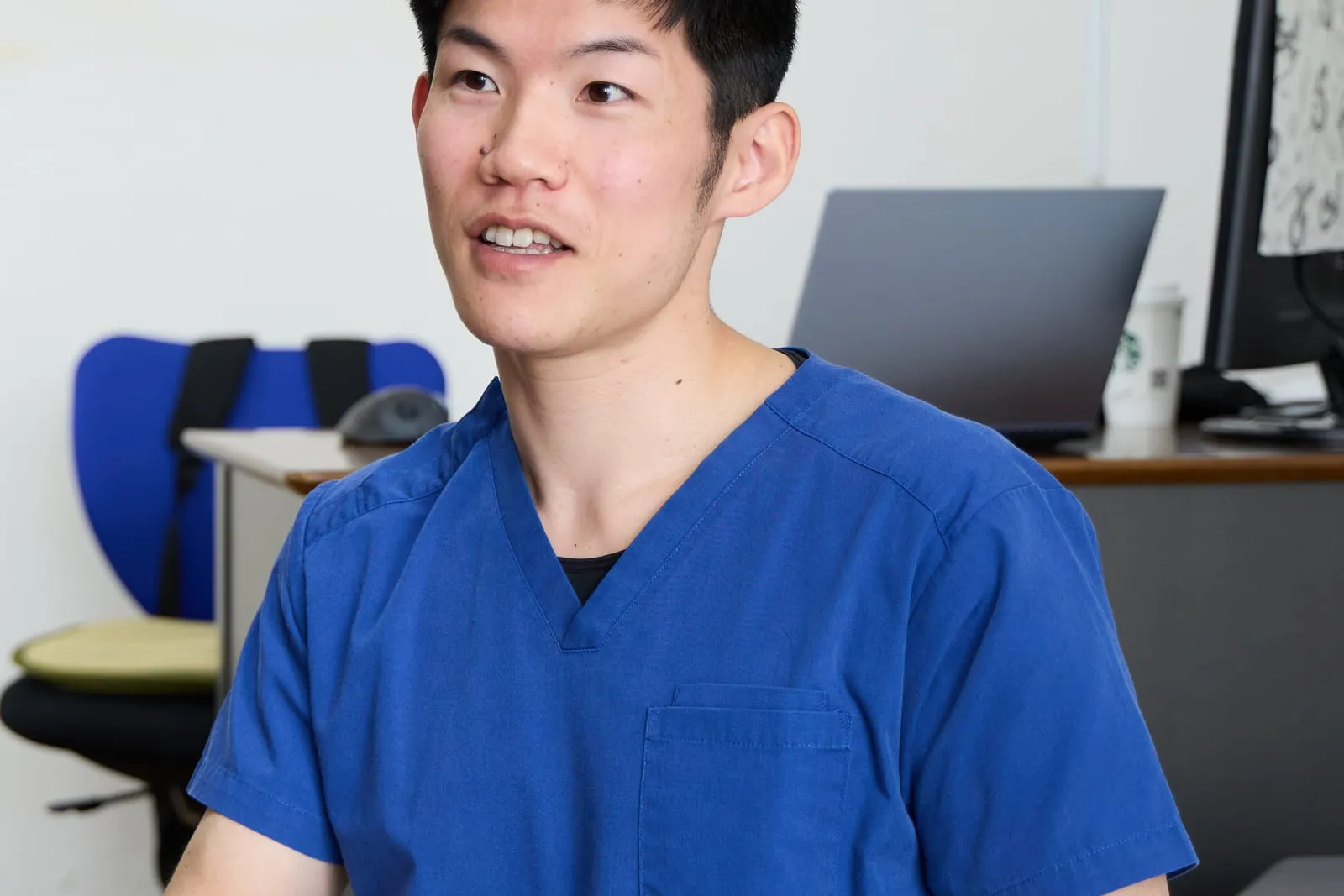 青い施術着を着た施術者 村上 優が、机とノートパソコンのある部屋で話しているところ。