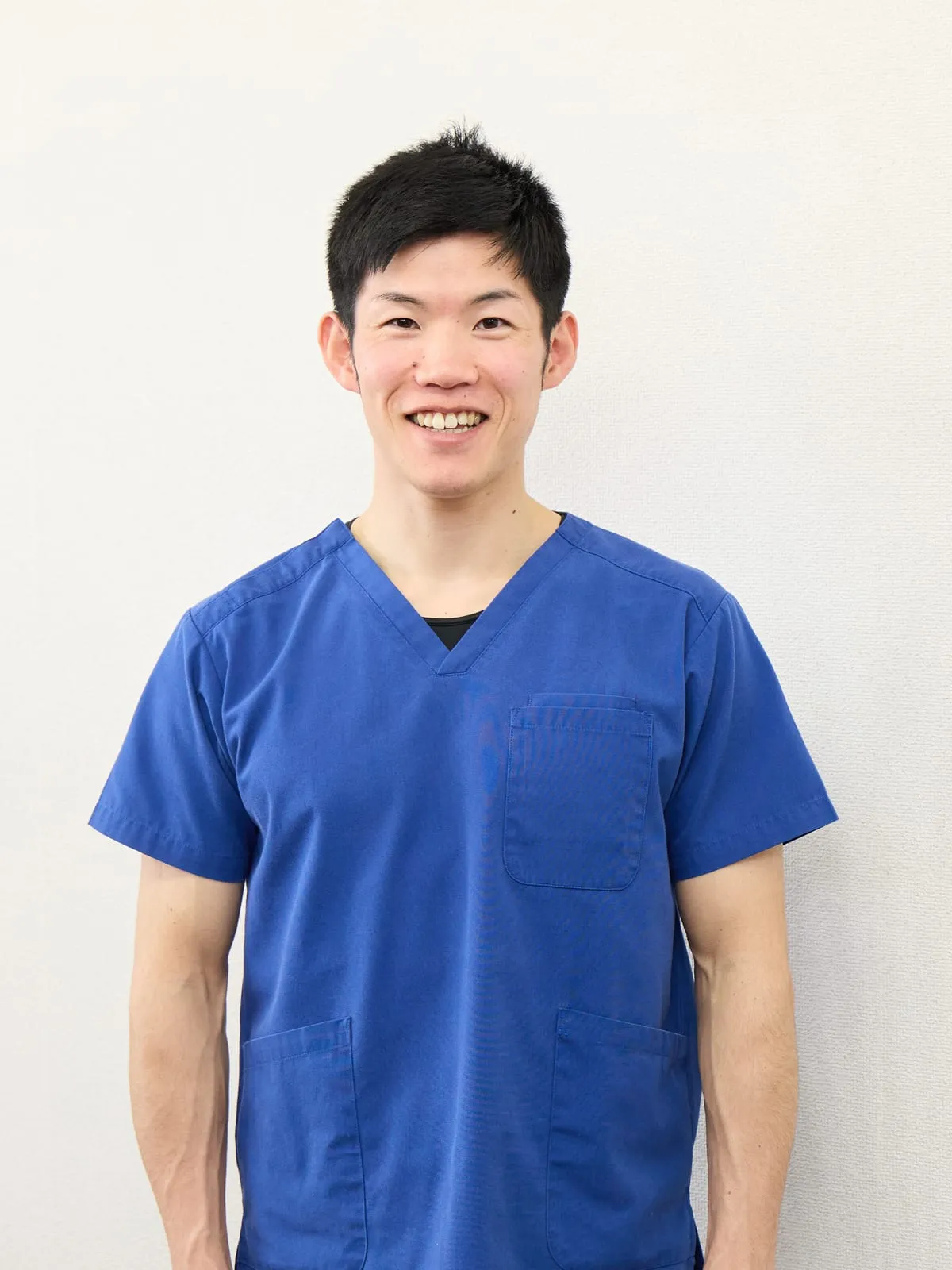 施術者 村上 優の写真。白い壁を背に、青い施術着を着て立っている。